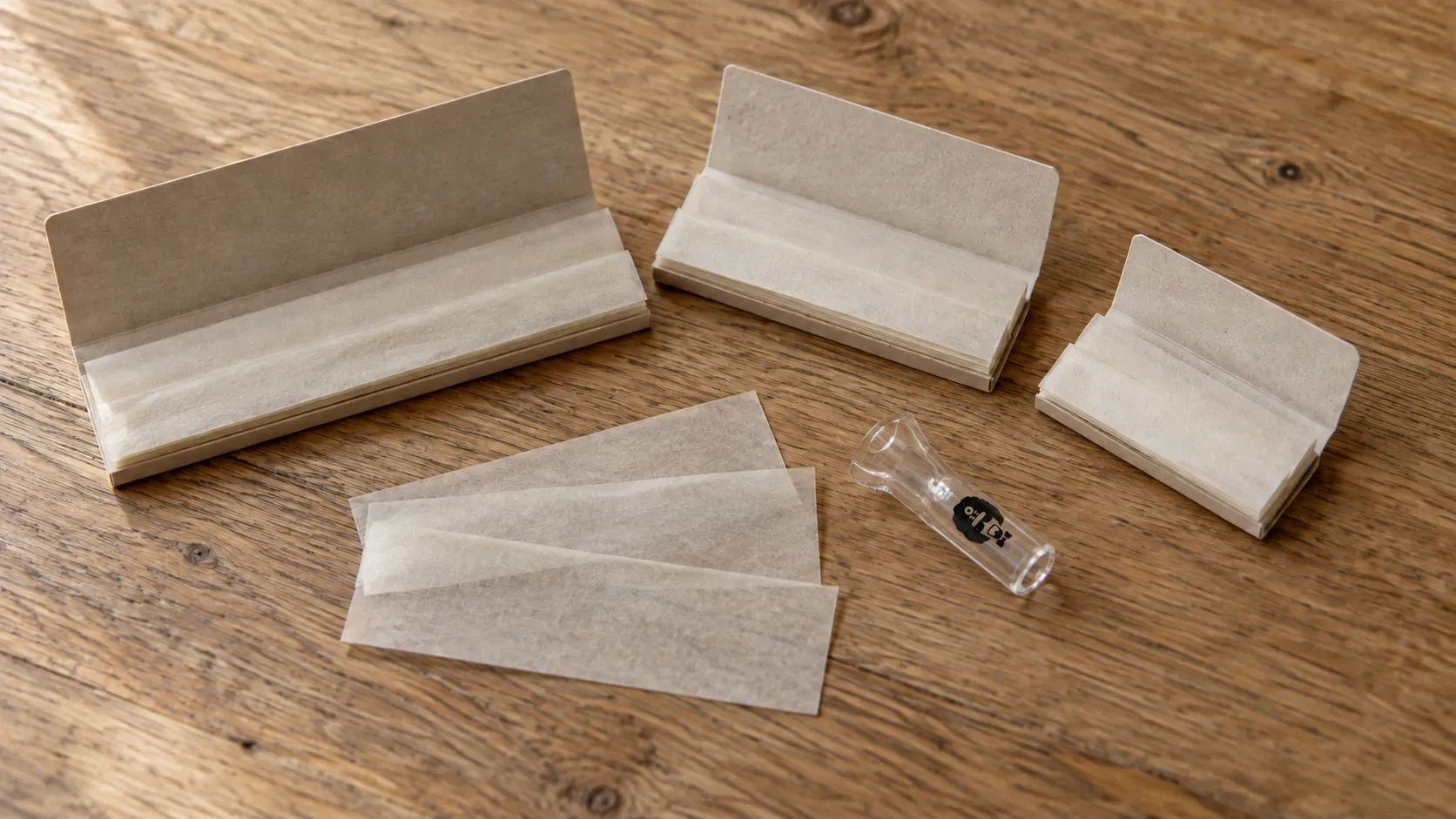 Several booklets of plain rolling papers in different widths laid out on a wooden table next to a clear glass tip
