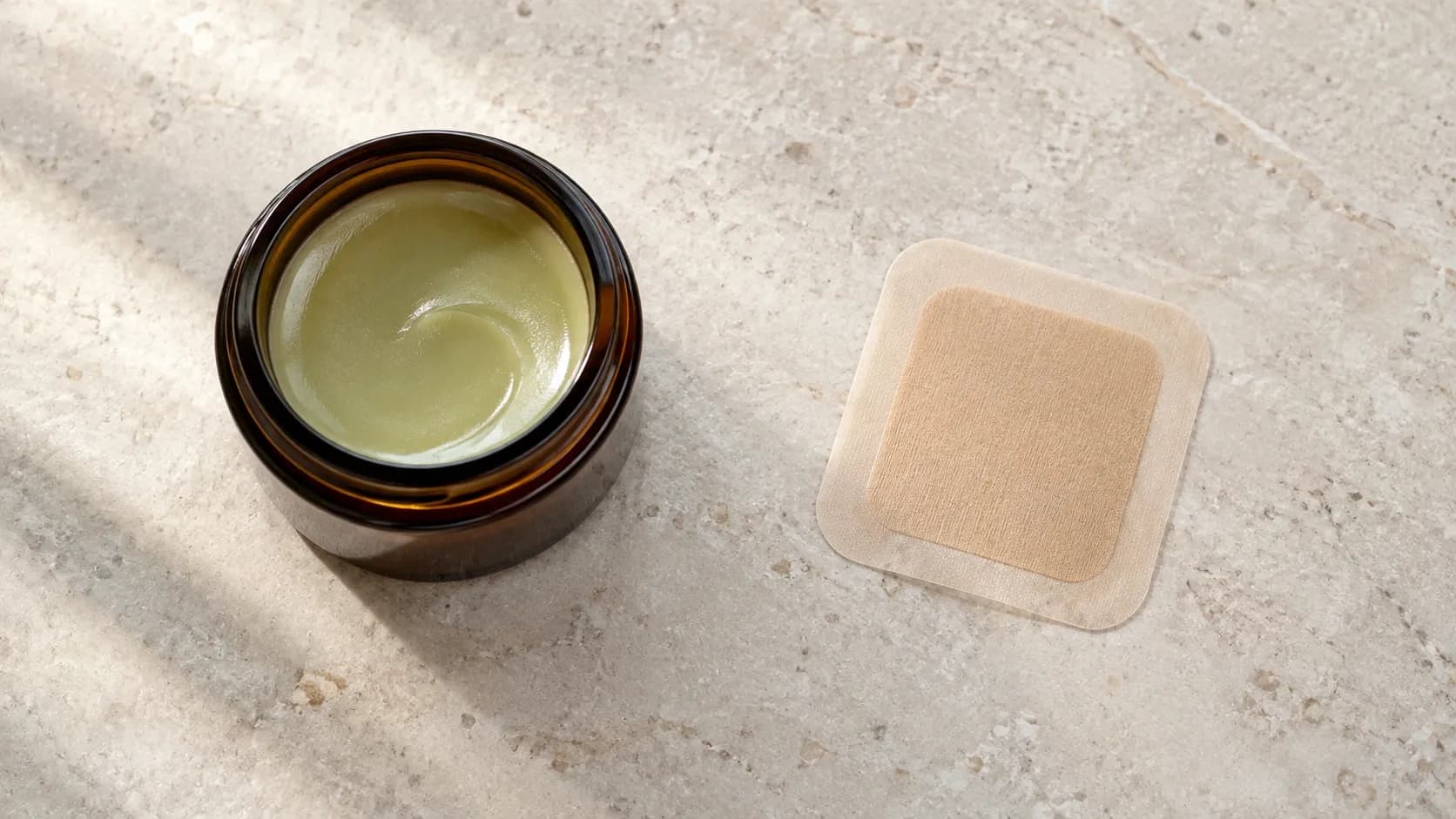 A jar of cannabis balm beside a transdermal patch on a clean pale surface